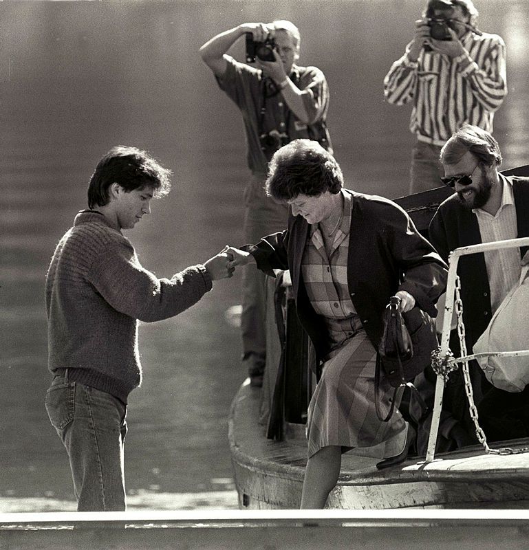 Black and white photo of a man holding a woman's hand on the way off a boat, while two people in the background are photographing. In the boat,  there's a man with sunglasses.