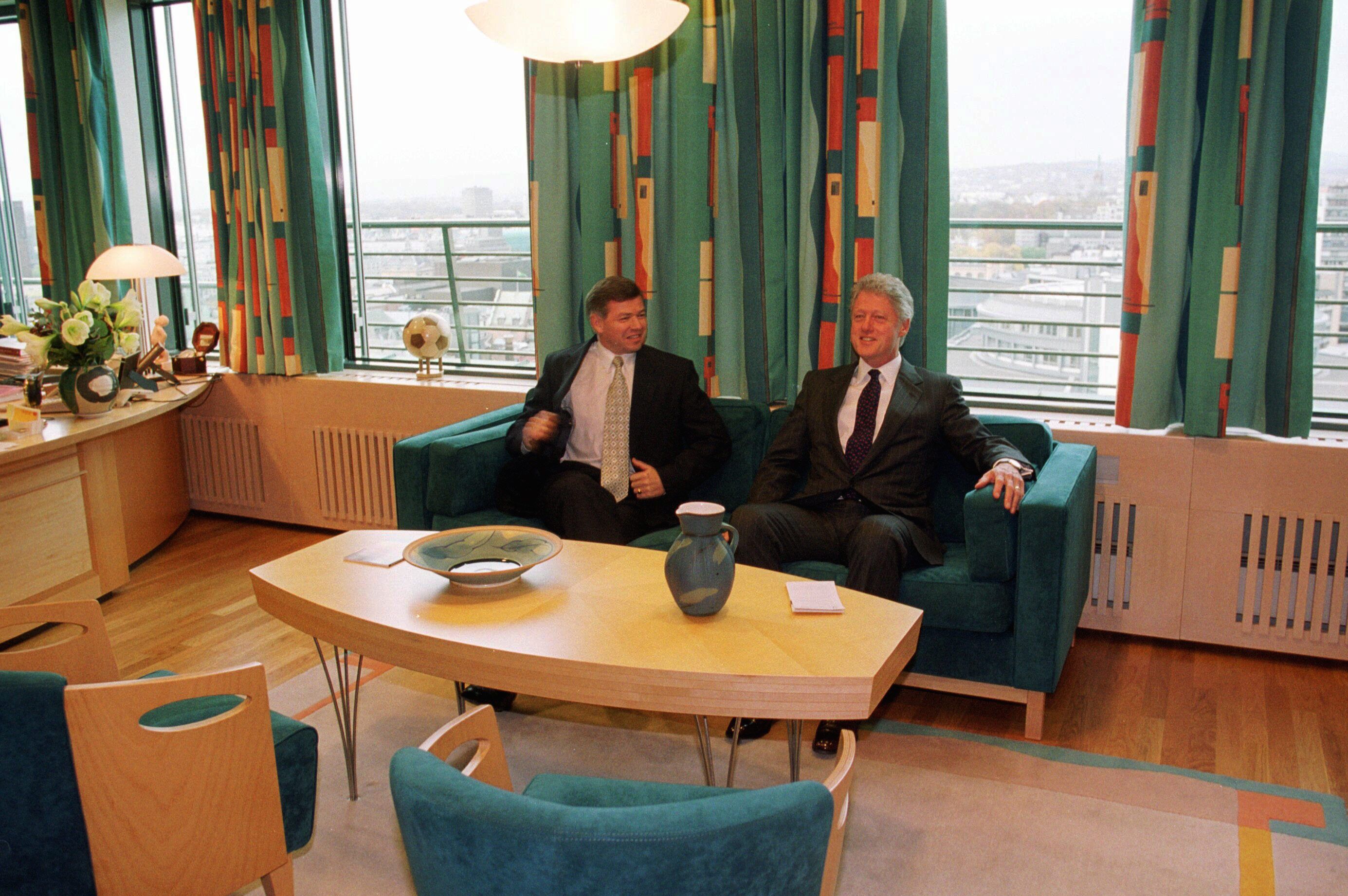 Two people in suits on a green sofa in an office with patterned curtains. Through the windows you look down on the cityscape.