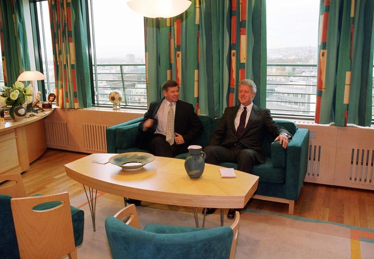 To dresskledde personer i grønn sofa i kontor med mønstrete gardiner. Gjennom vinduene ser man ned på bylandskap.  