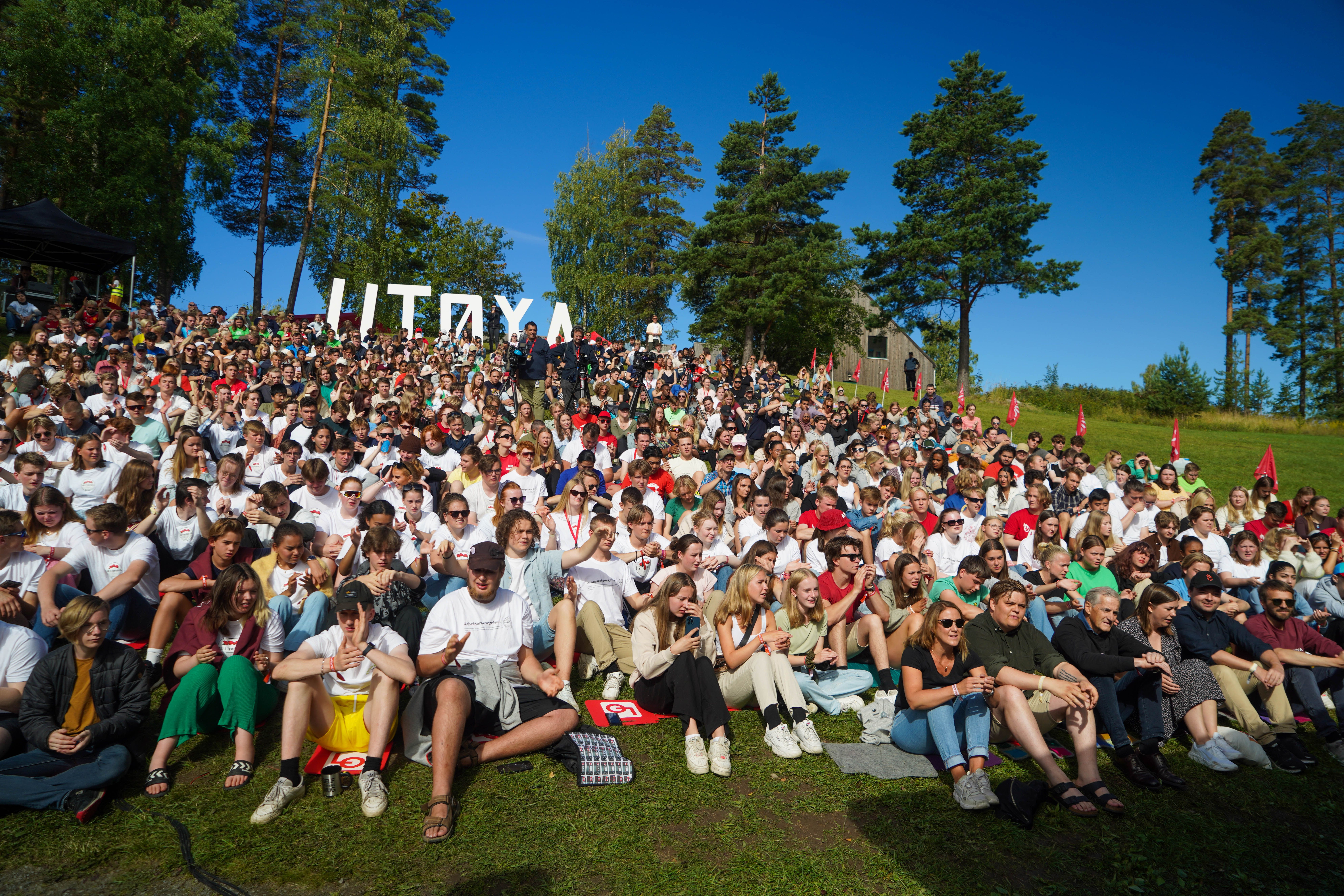Crowd sitting on grassy hill with trees and bushes. In the background large figurative letters form "UTØYA"