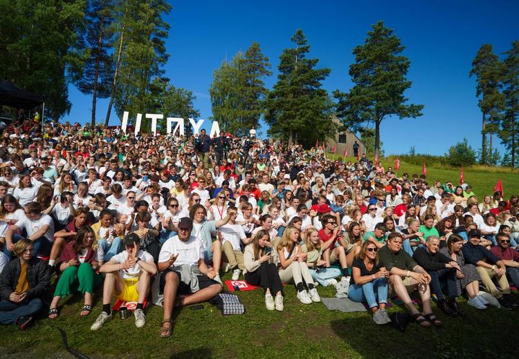 Crowd sitting on grassy hill with trees and bushes. In the background large figurative letters form "UTØYA"