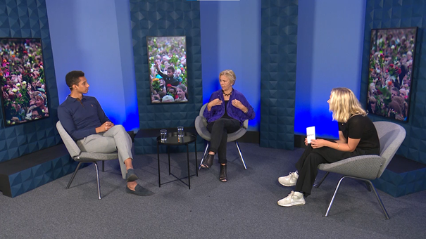 Three people are sitting and talking to each other in front of a blue background with three large pictures of people with roses.