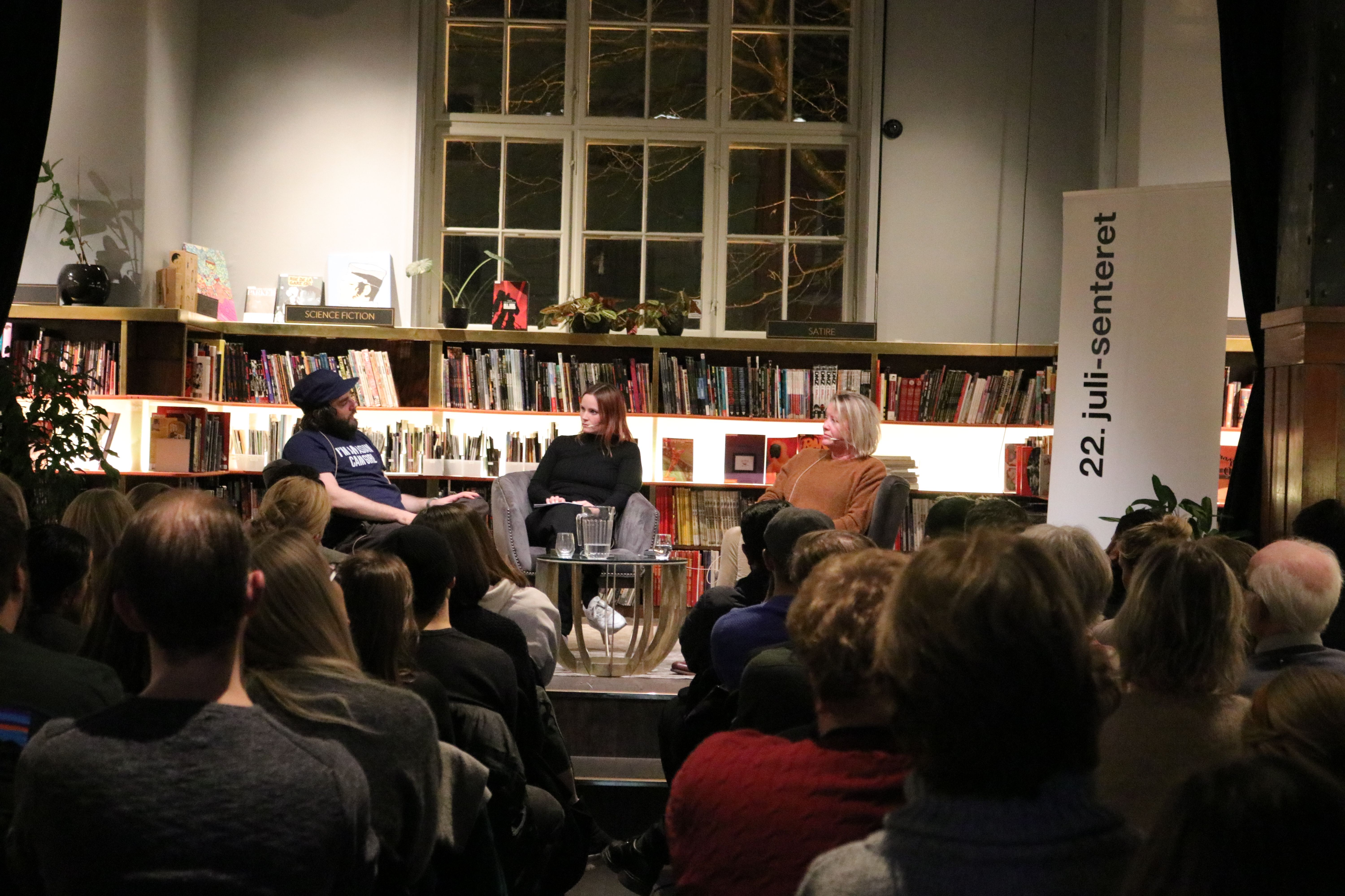 Three people sit in a scene. In the background a large bookshelf with books. The audience sits in front of the stage.