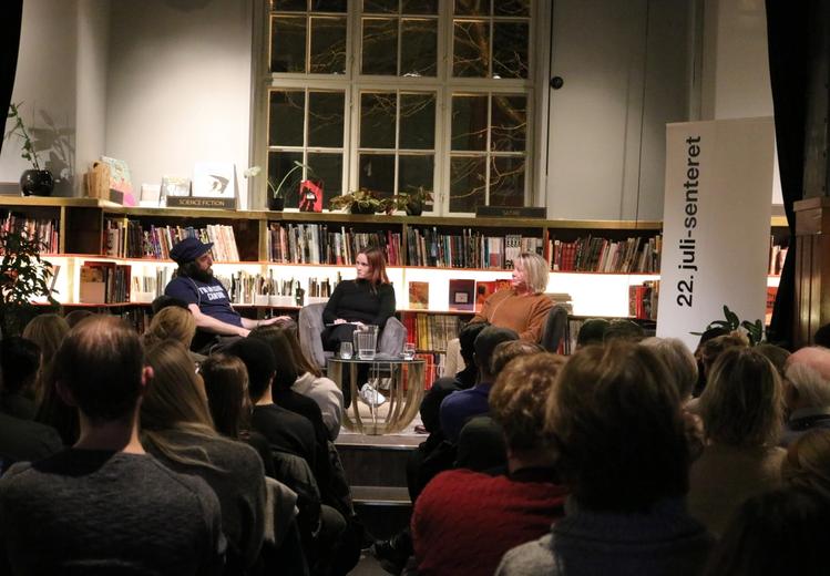 Three people sit in a scene. In the background a large bookshelf with books. The audience sits in front of the stage.