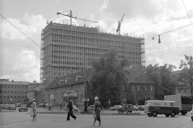 Bilde i svart-hvitt av stilasomringet bygg og heisekraner fremfor åpen plass med rundkjøring. I forgrunnen er tre spaserende personer og flere vintagebiler er synlige.