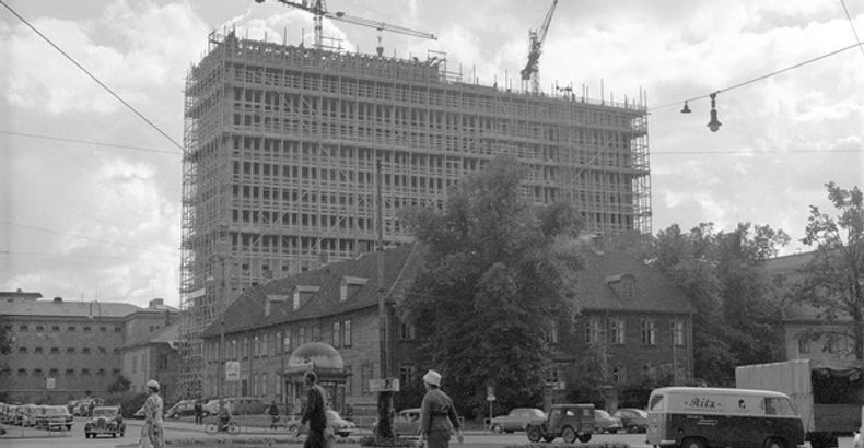 Black and white photo of a building surrounded by lift cranes, in front of an open space with a roundabout. In the foreground, three people walking and several vintage cars are visible.