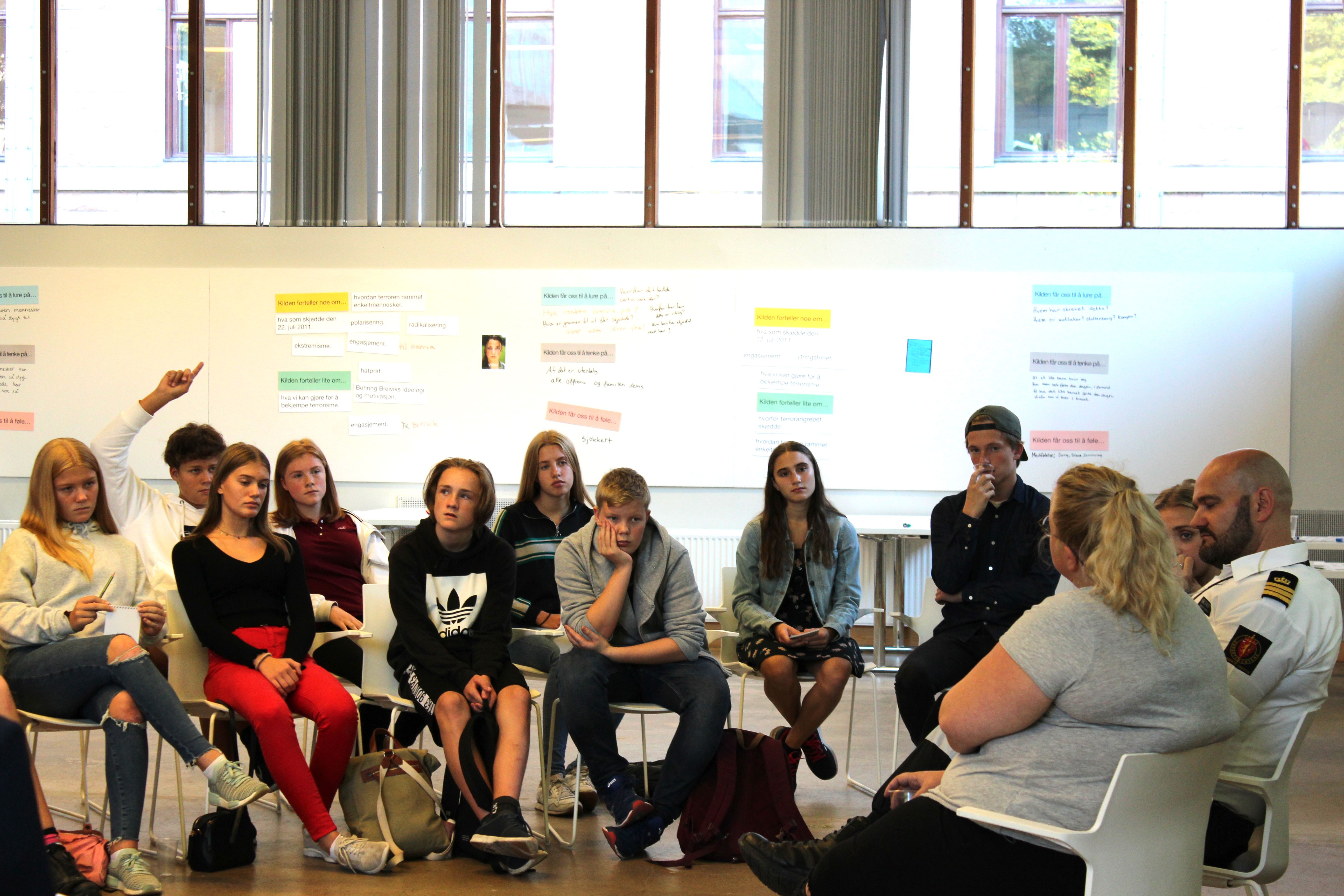 Pupils sitting on rows watching two people talking. One of the people Talking wears a uniform. The room they are sitting in is white and in the background there is a whiteboard with written text and images. 