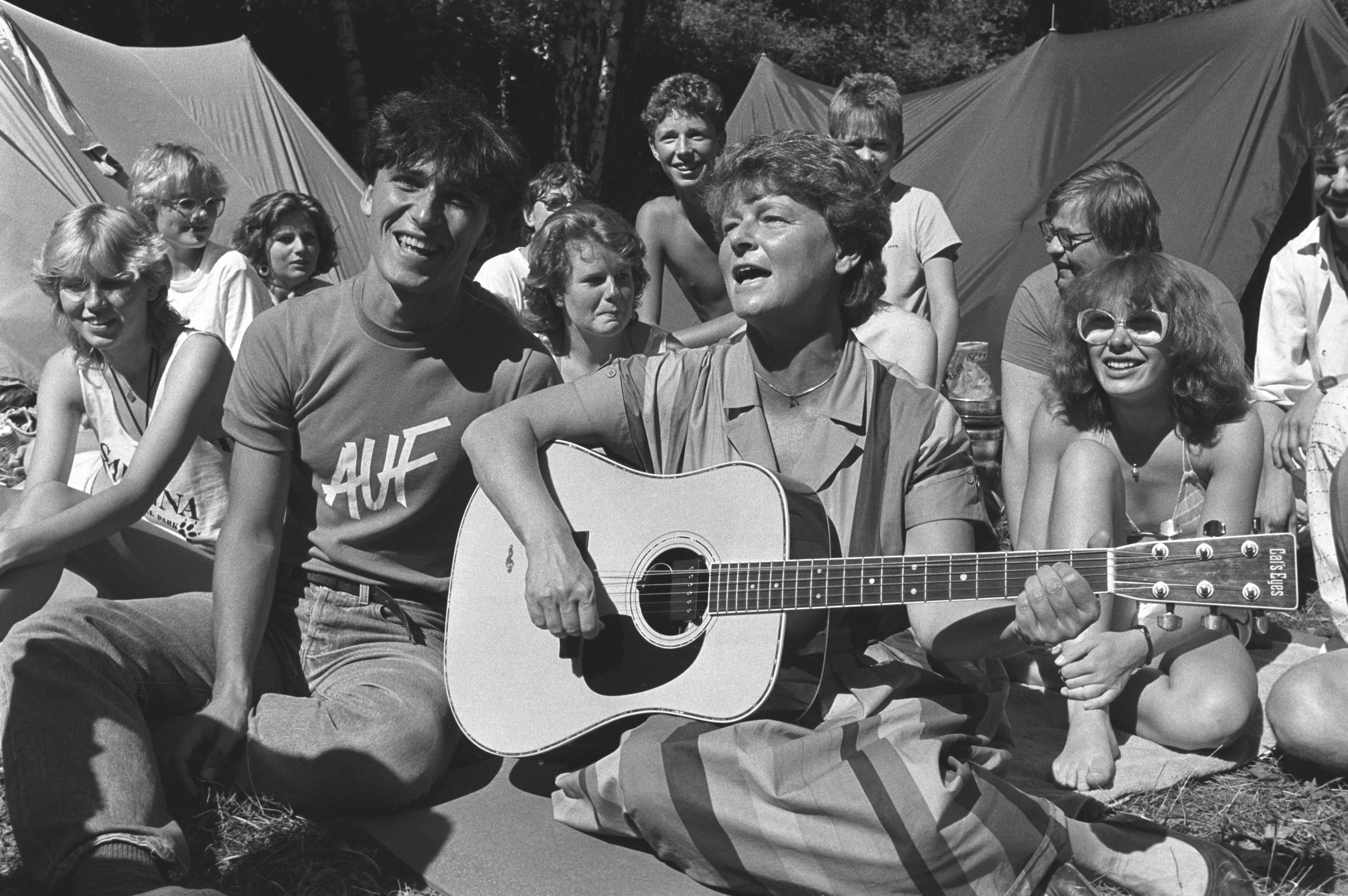 Black and white photo of woman with guitar and group of people sitting in the grass. A man sitting on the left of the woman has a t-shirt with the inscription "AUF". Two tents and trees can be seen in the background.