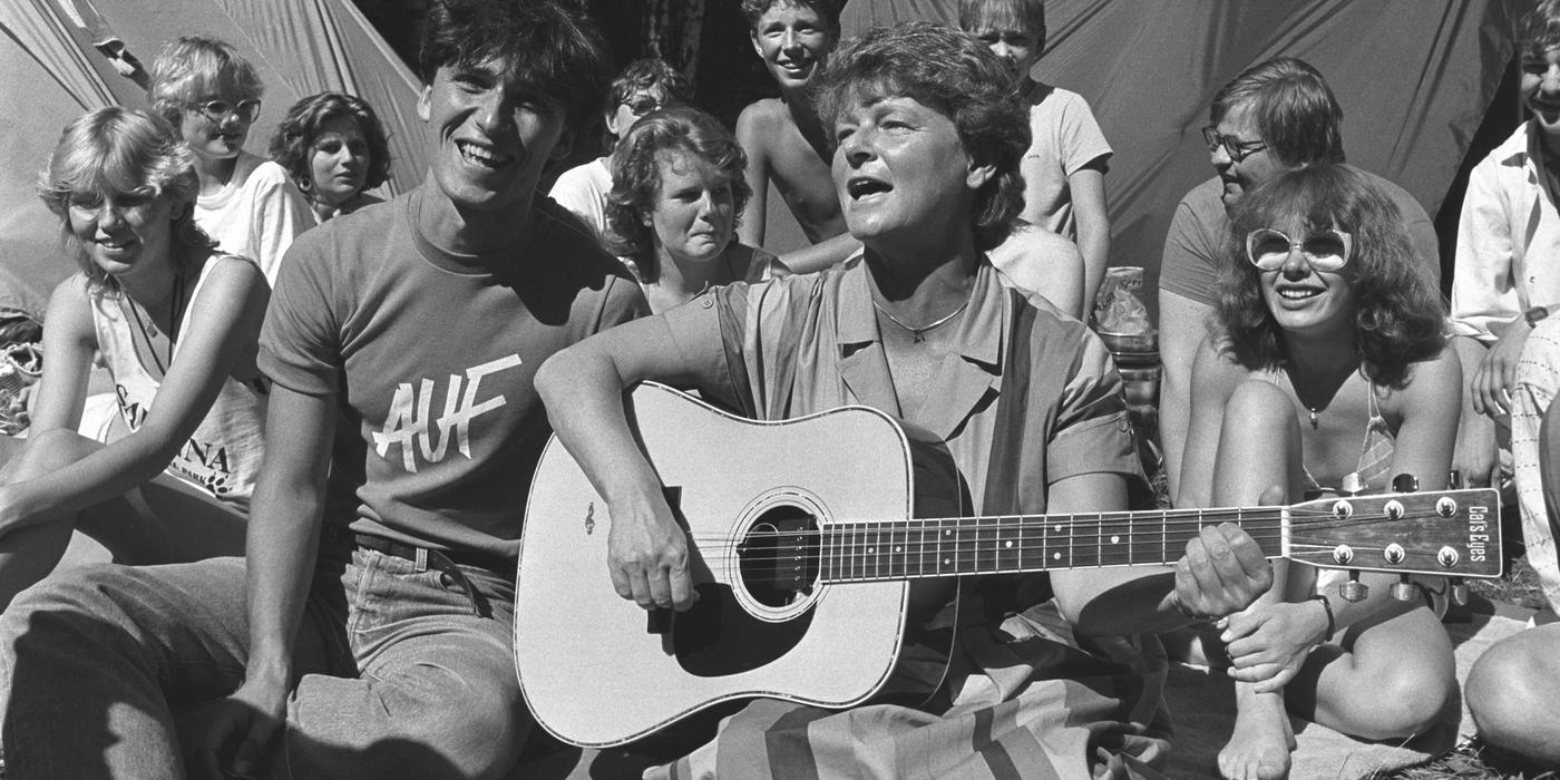 Black and white photo of woman with guitar and group of people sitting in the grass. A man sitting on the left of the woman has a t-shirt with the inscription "AUF". Two tents and trees can be seen in the background.
