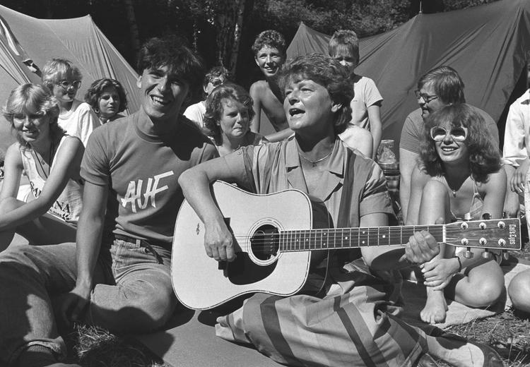 Bilde i svart-hvitt av kvinne med gitar og samling av mennesker sitter i gresset. Mann har t-skjorte med påskrift "AUF". I bakgrunnen synes to telt og trær.  
