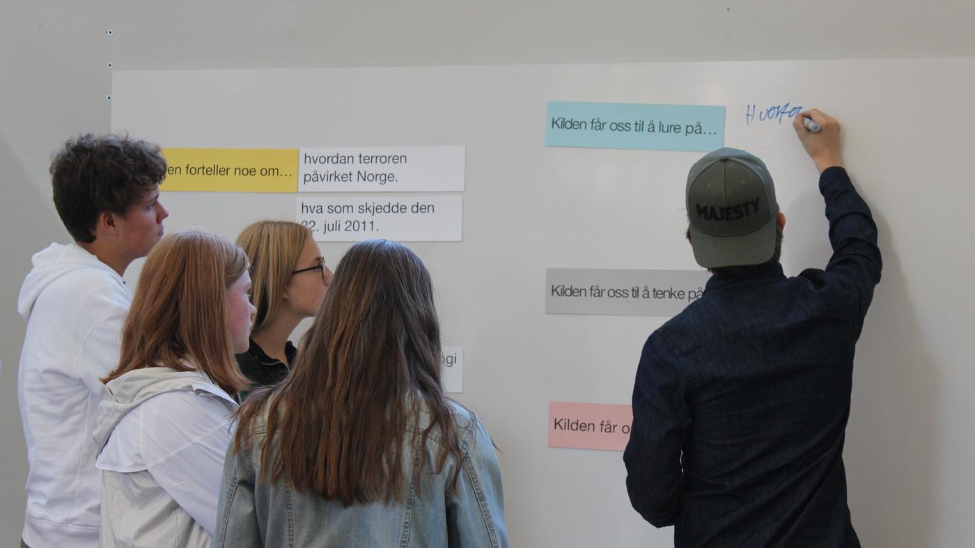  Four students stand in a group, one student wearing a dark shirt and gray cap draws on the blackboard. Six pieces of paper in different colors with text: "(..) tells about", "How terror affected Norway", "The source makes us wonder", "what happened on 22 July 2011", "The source makes us to think about.." "The source gets (..)"