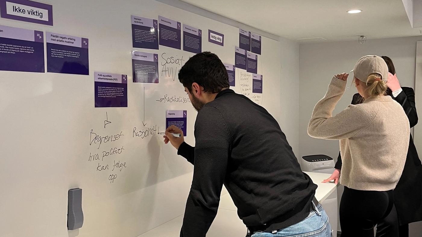 Three people working around a white magnetic board with purple card on it. The cards have a title and a text below. One of the persons is writing on the board with a marker.