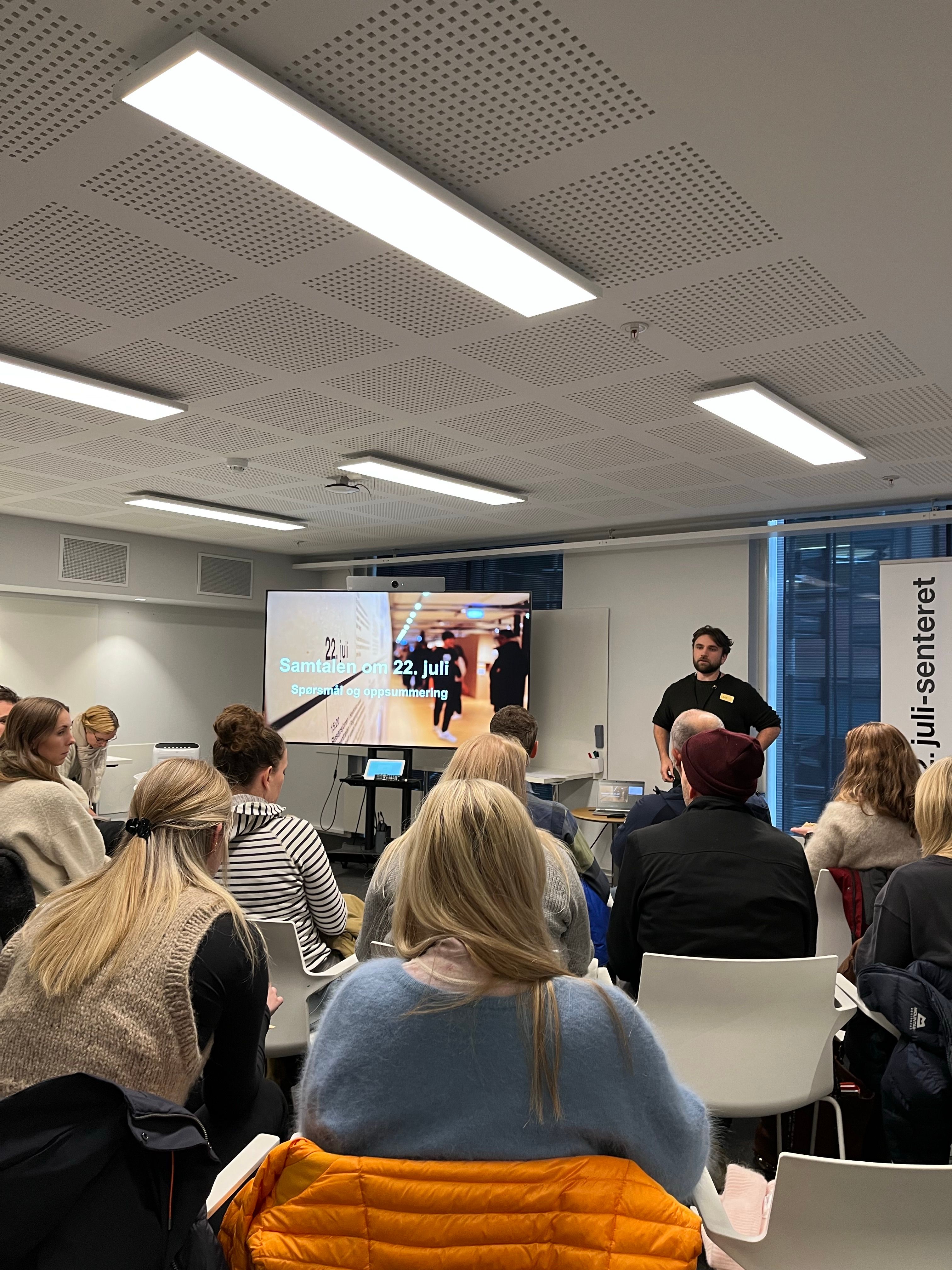 En gruppe sitter på hvite stoler. De ses bakfra mens de ser på en man med sort t-skjorte som snakker. Ved siden av mannen ses et stort skjerm med et bilde av en utstilling og tittelen Samtalen om 22. juli. Spørsmål og oppsummering.