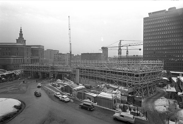 Black-and-white image of building with scaffolding and cranes in front of motorway. More vintage cars. Snow appears on the ground.
