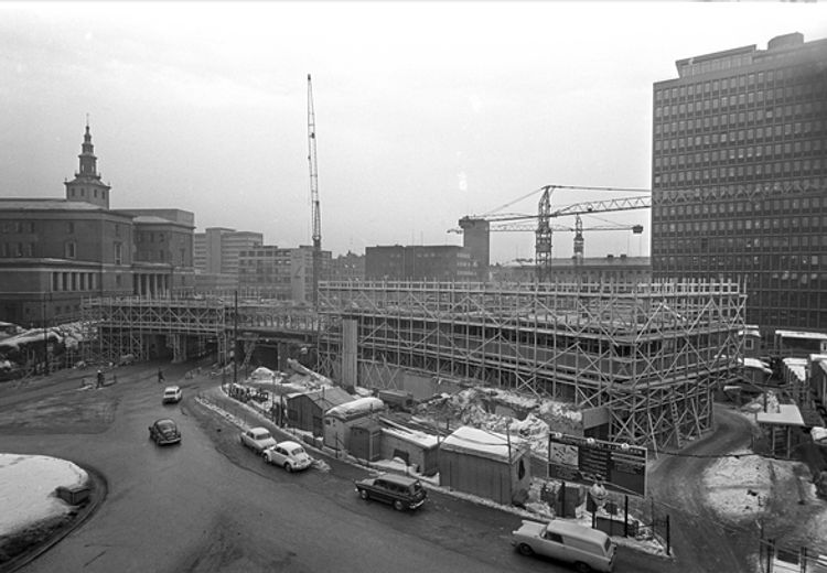 Black-and-white image of building with scaffolding and cranes in front of motorway. More vintage cars. Snow appears on the ground.