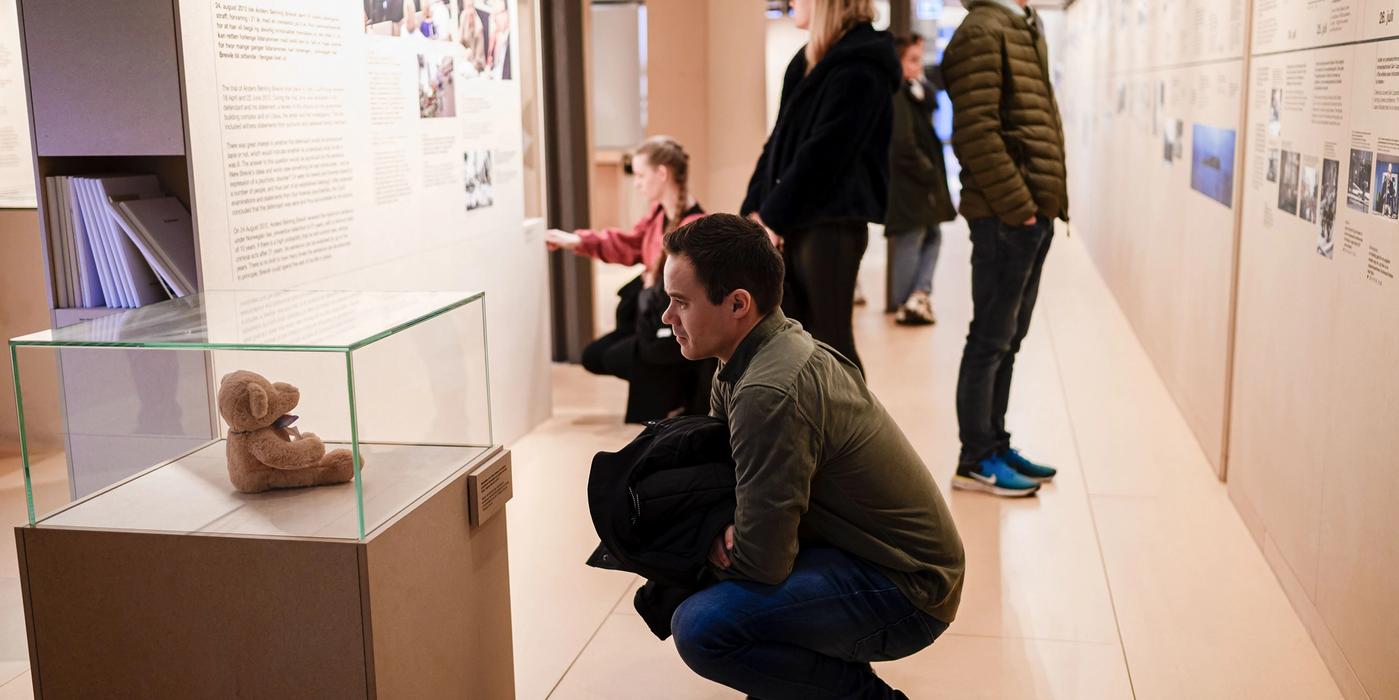 Five visitors look at an exhibition. In the foreground a man is squatting looking at a teddy bear that is inside a display case.