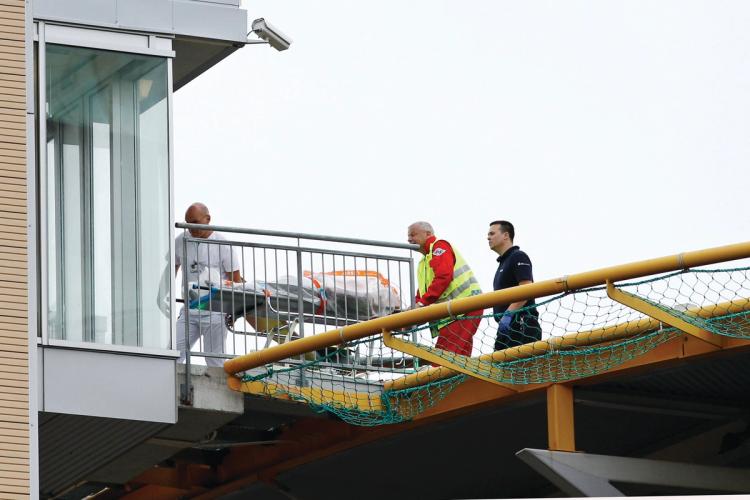 Three people carry a stretcher on a helicopter deck. Build in front of the image. Gray sky in the background.