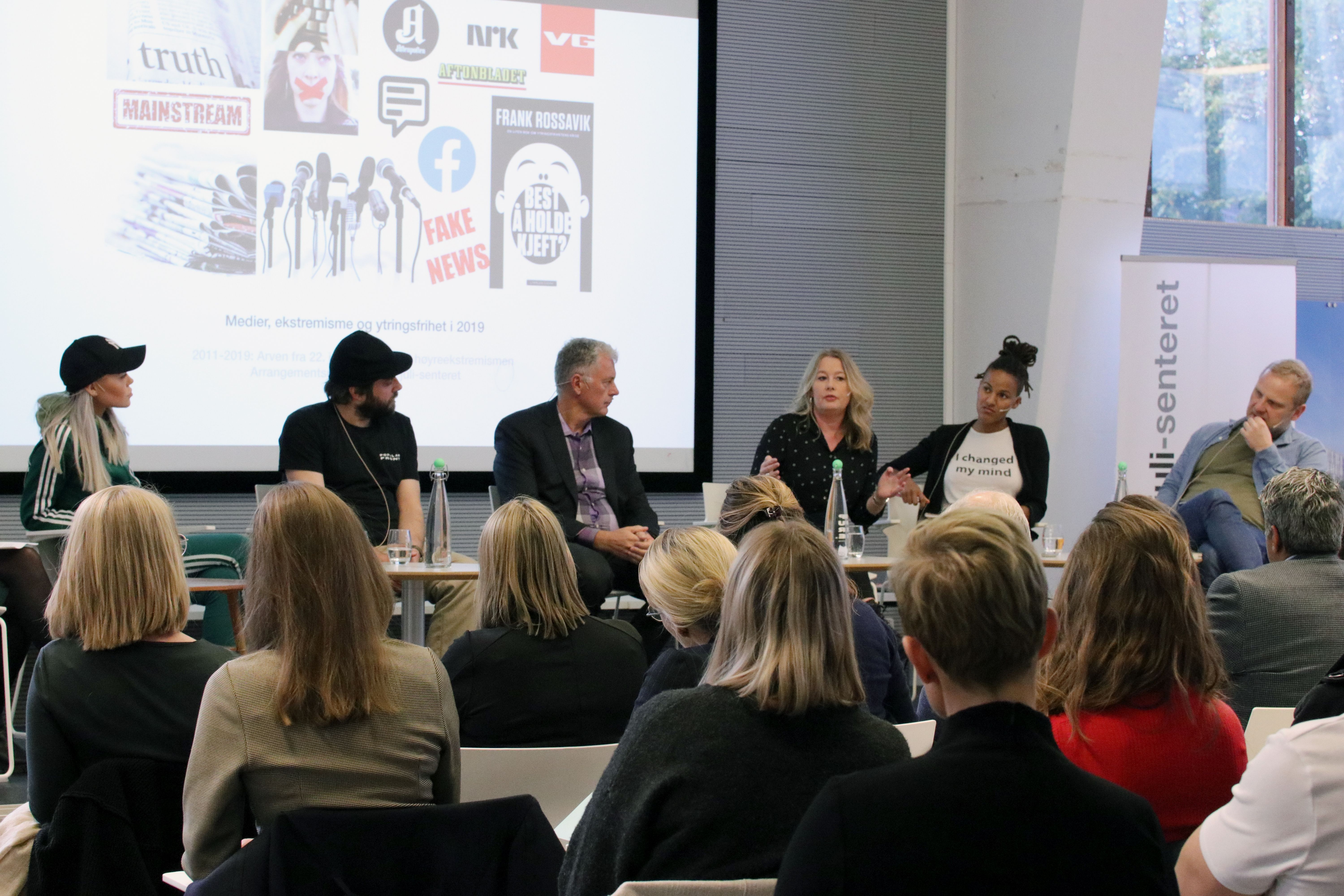 Six people sit in front of a large screen with indistinct figures. A woman among the six speaks. An audience sits and listens.