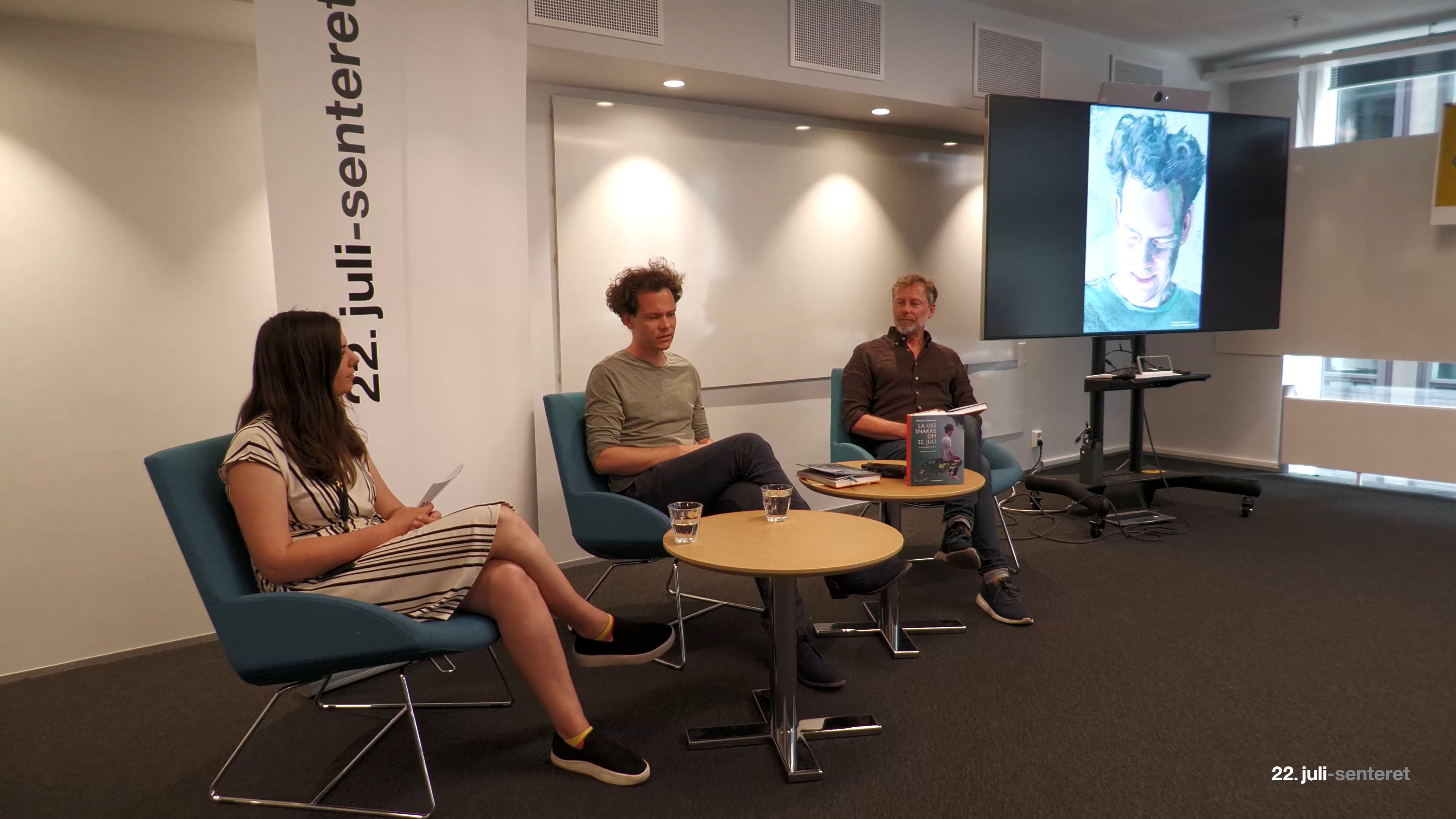 Three people sit in a panel discussion. To the right is a screen with an indistinct portrait, and behind the forehead moldings a roll-up with the 22 July centre's logo.