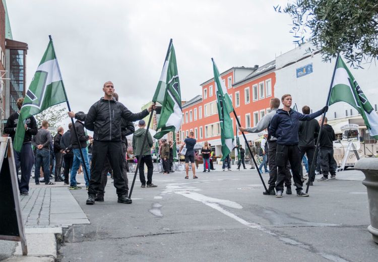 Several men is standing with green and white flags. They are standing on a road. There are buildings in the background.