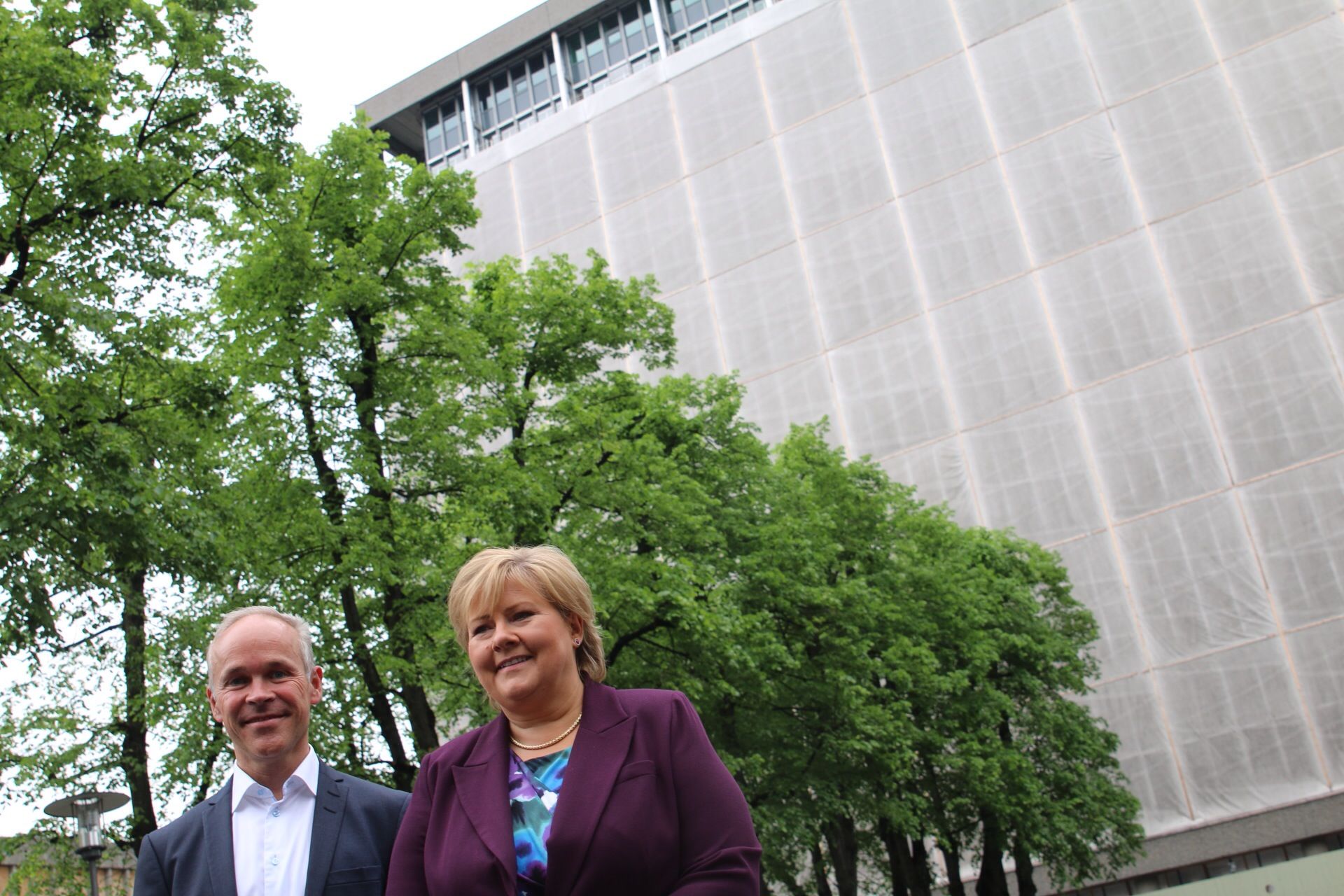  Two people in the front in the picture. One dressed in a blue suit with short hair. One dressed in a purple suit jacket with a blue colored blouse and blond short hair. Avenue with trees and a tall building with a covered facade in the background.