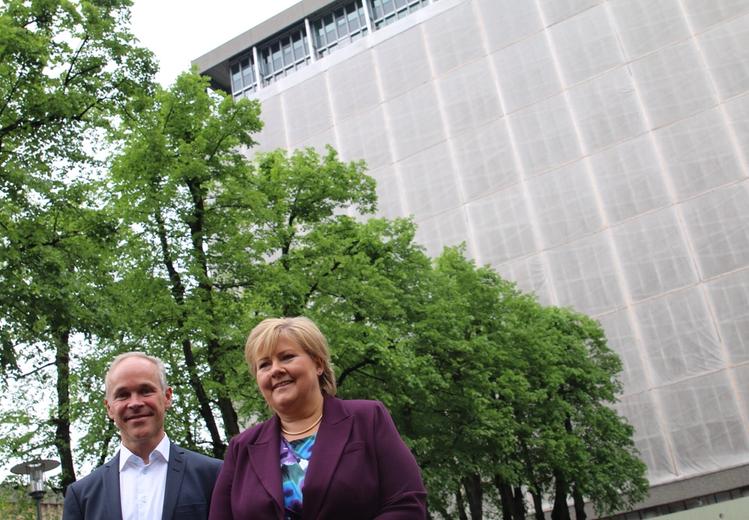  Two people in the front in the picture. One dressed in a blue suit with short hair. One dressed in a purple suit jacket with a blue colored blouse and blond short hair. Avenue with trees and a tall building with a covered facade in the background.