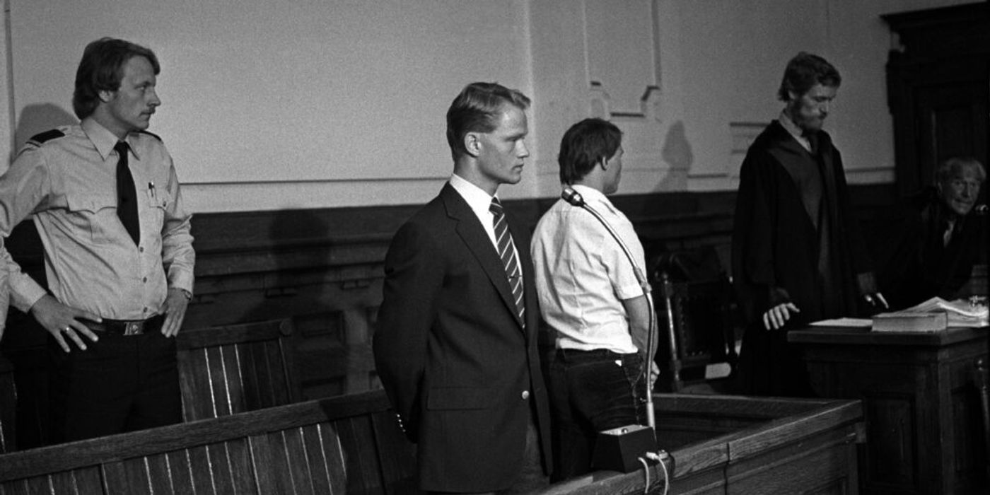 A man stands in a suit with a microphone in front of him. A guard stands behind him. A man in a judge's robe stands in the background. The interior is in wood. The picture is in black and white.