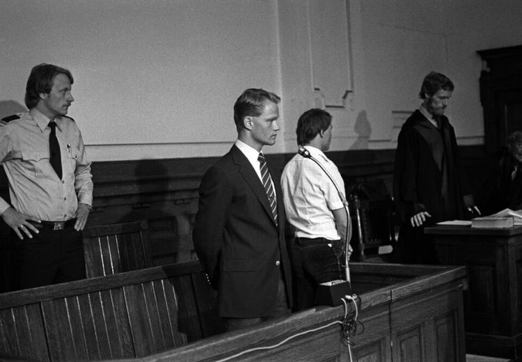 A man stands in a suit with a microphone in front of him. A guard stands behind him. A man in a judge's robe stands in the background. The interior is in wood. The picture is in black and white.