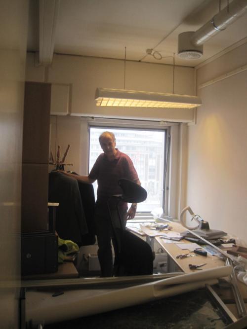 Foto av en mann som står midt i et ødelagt kontor, med et vindu uten glass bak seg.