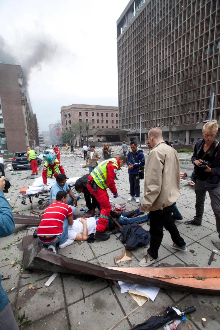 Photo. People in front of buildings with smoke in city landscape. In the front, a man lying down receiving help from other persons gathering around him.