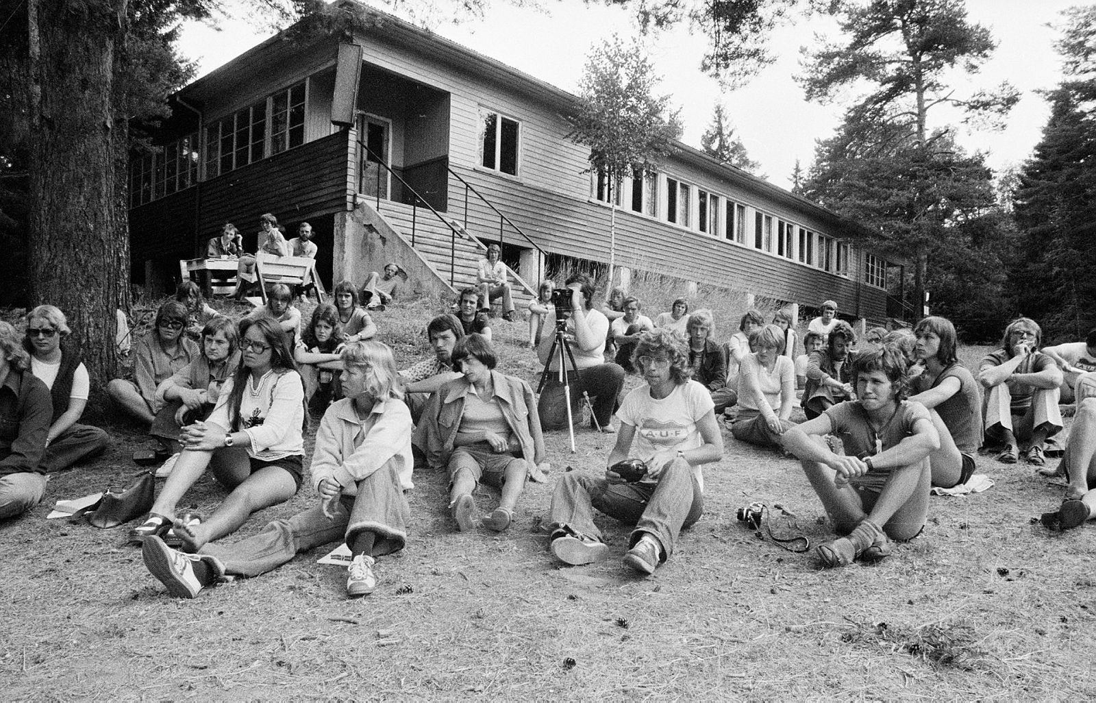 Black and white photo of people sitting outside a low wooden house with a staircase. In front of the staircase, a person with a camera and a tripod. Three people are sitting at a picnic table.