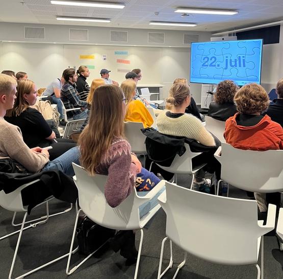 People gathered in a classroom sitting on white chairs, looking away from us at a screen with the text "22 July"