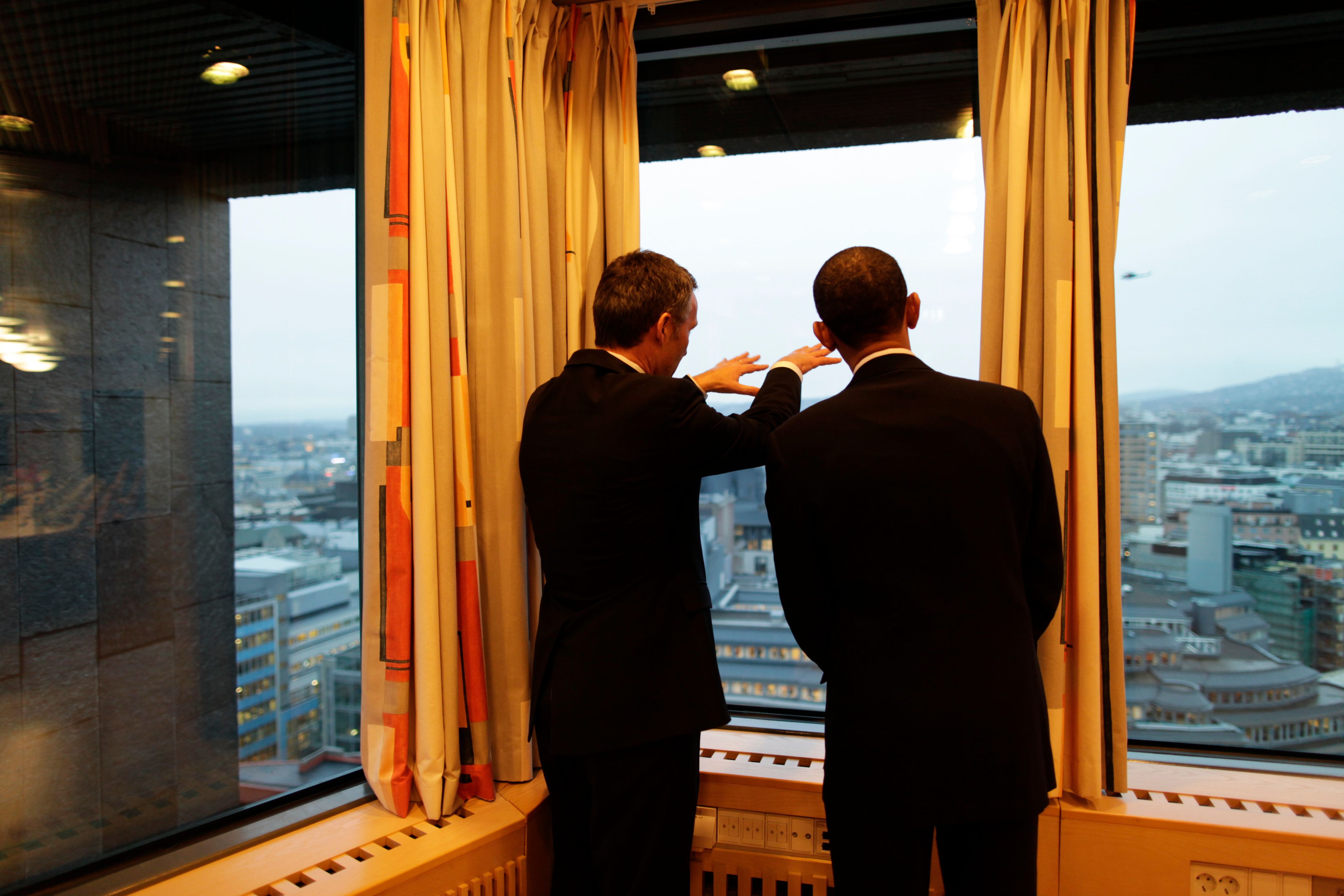 Photo of two men seen from behind who are looking out a window of a top floor. The man on the left is pointing.