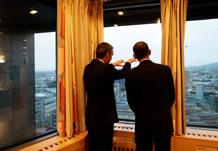 Photo of two men seen from behind who are looking out a window of a top floor. The man on the left is pointing.