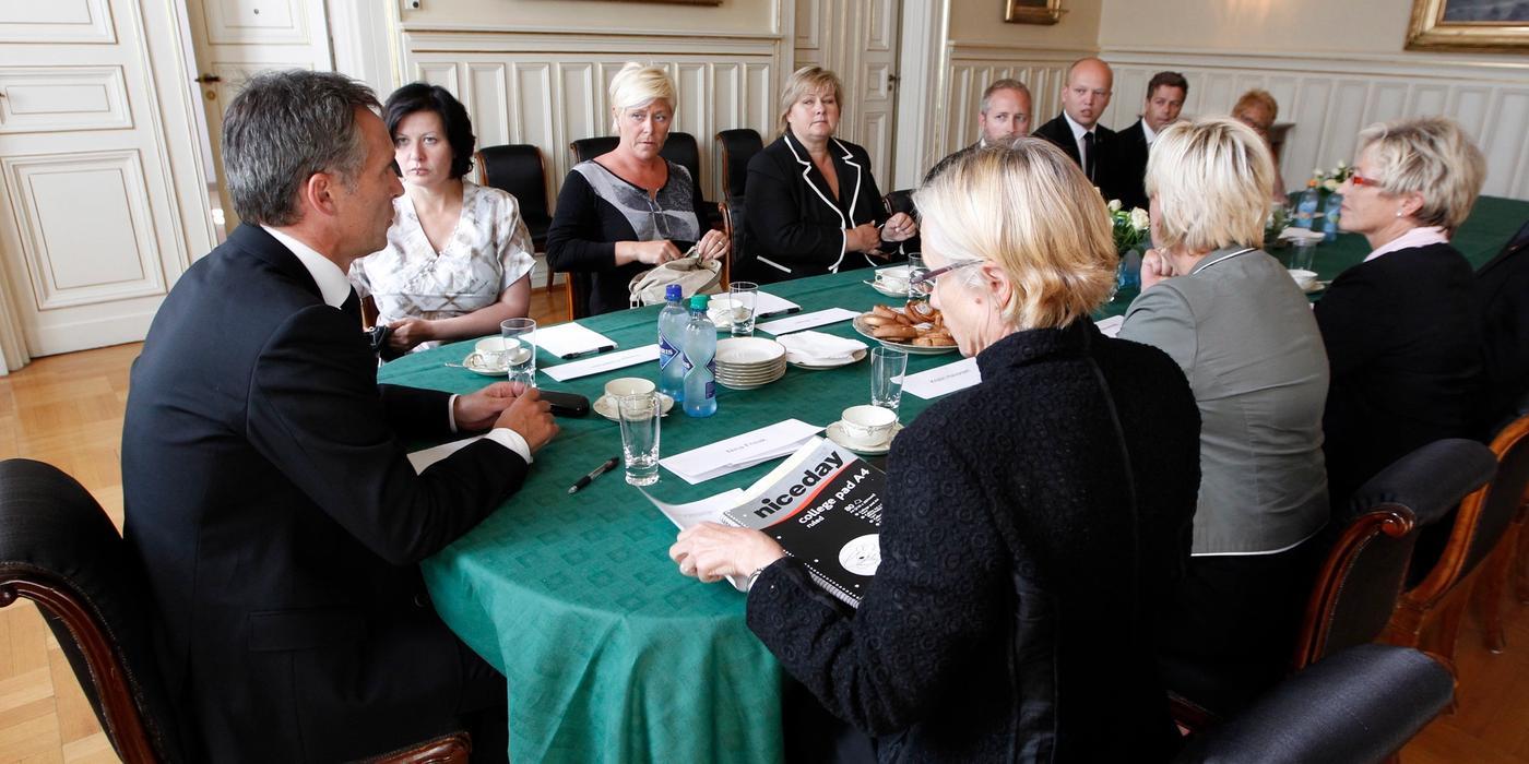 Rundt et bord med grønn duk sitter elleve mennesker. Ti av dem lytter til den som sitter på toppen av bordet. Vedkommende har på seg dress. Han var statsminister i Norge i 2011. Fotografi. 