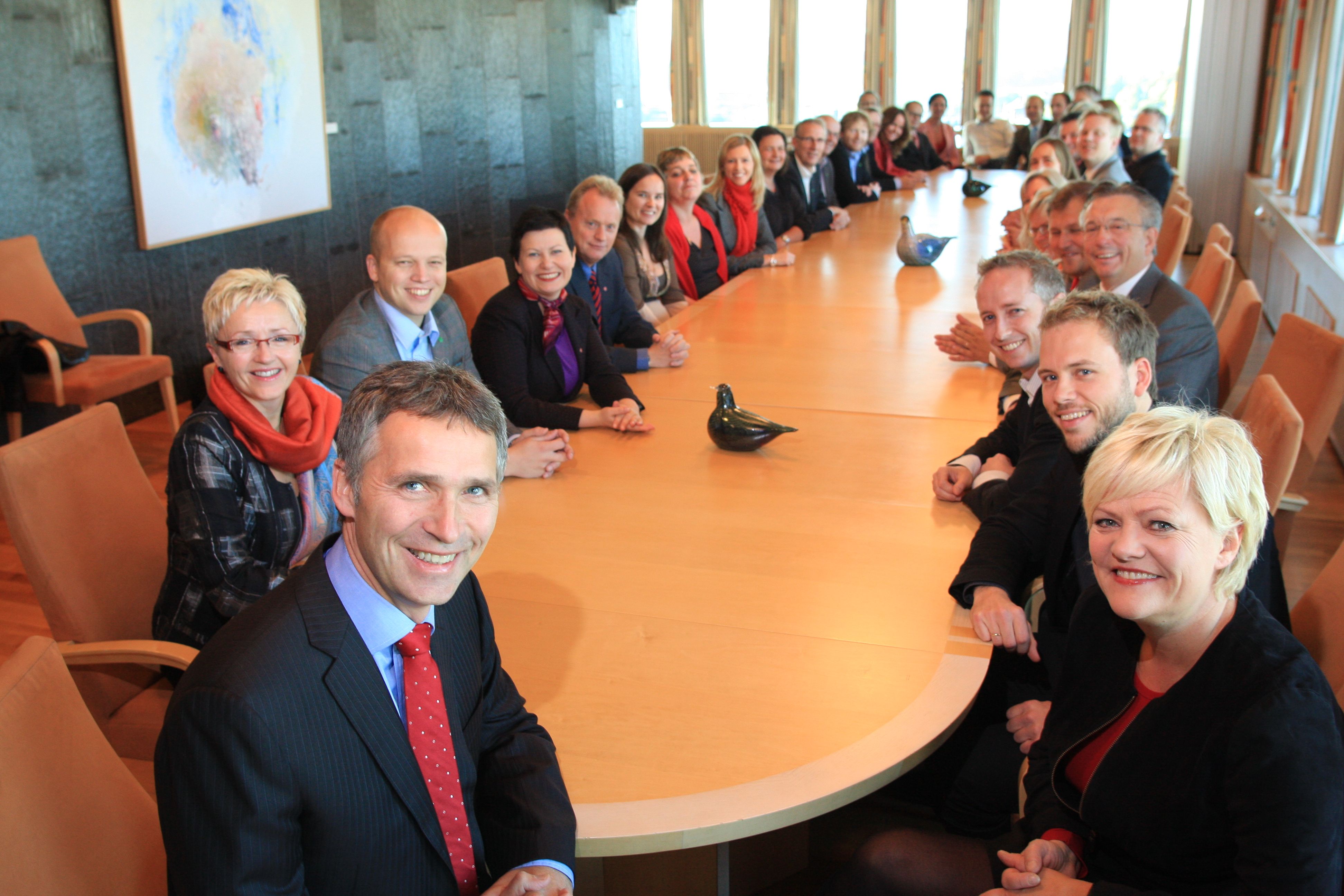 Many men and women are sitting around a large wooden table. They are smiling and is nicely dressed. Several large windows. A wall of slate stone.