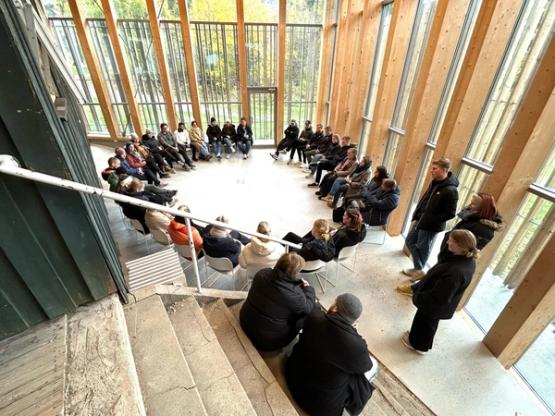 Several people sit on chairs, one side has a black wooden wall with windows and a clean glass side with light wood.
