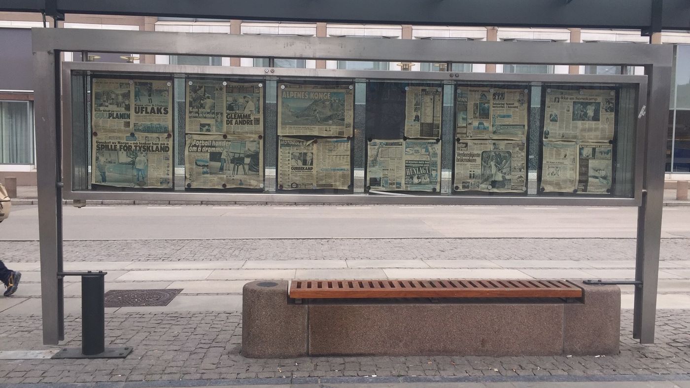 Stengulv. Busstopp i stål. Benk under i sten med tre. Avisforsider med uklar tekst.