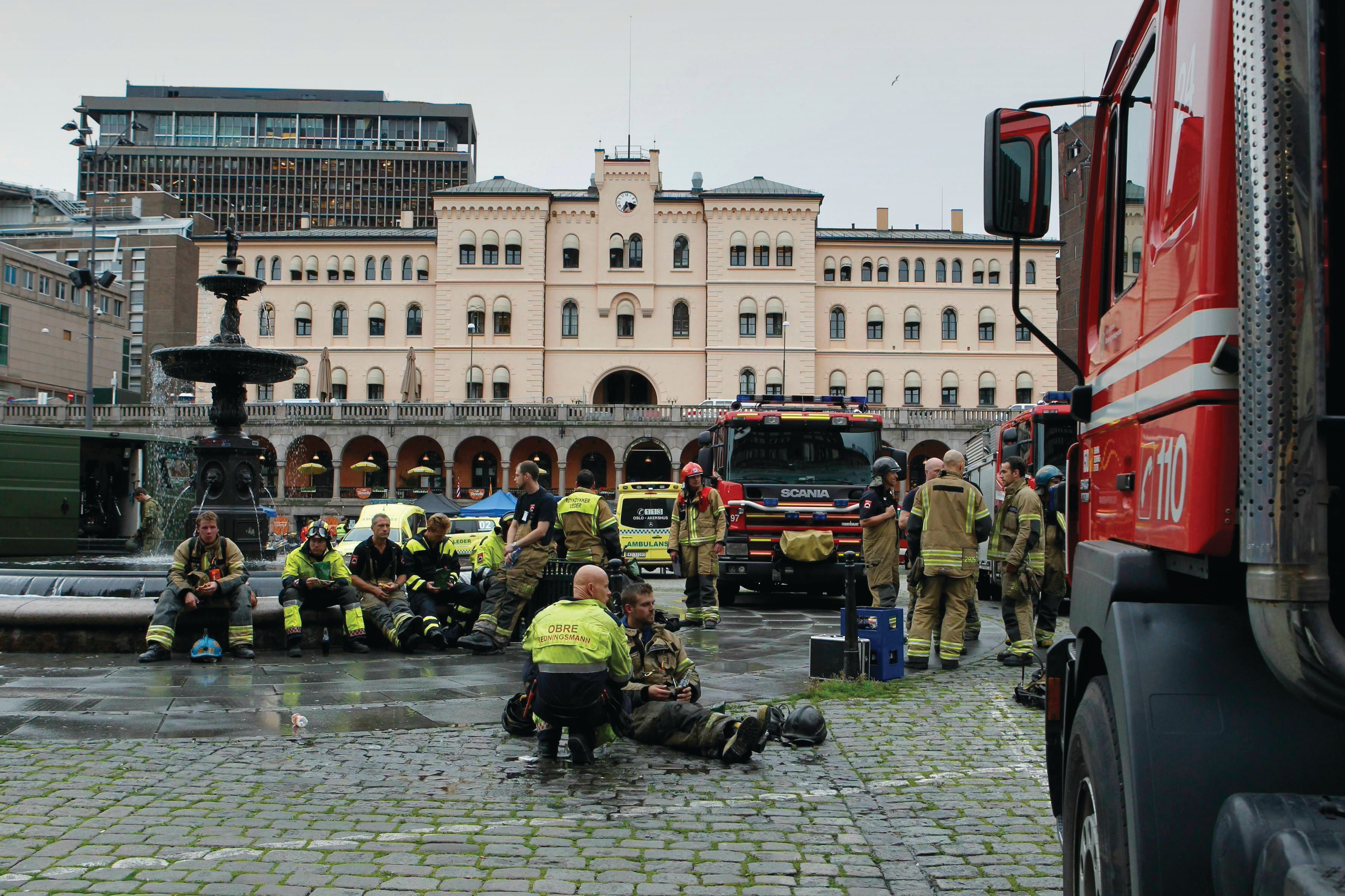 Red fire engine on cobblestones in the foreground. Twelve people in uniform. Fountain. Urban landscape. Building in pink and a tall building in the background