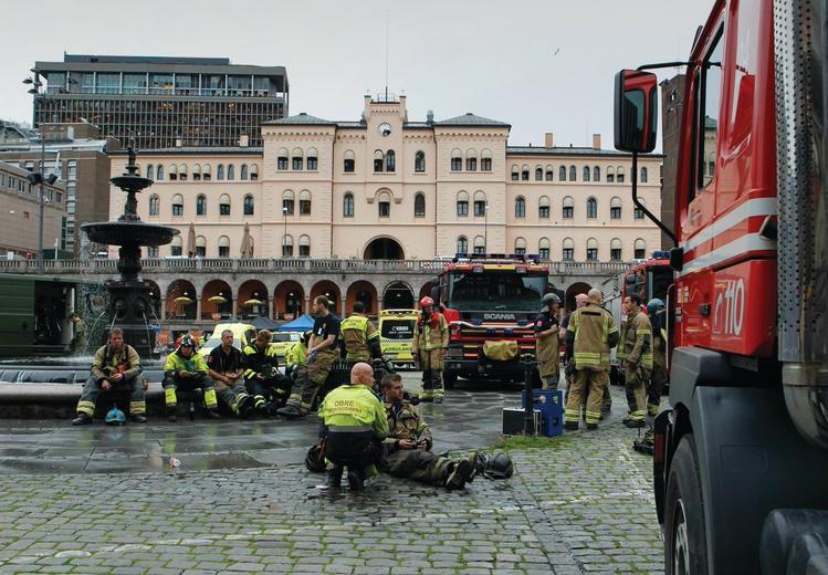 Red fire engine on cobblestones in the foreground. Twelve people in uniform. Fountain. Urban landscape. Building in pink and a tall building in the background