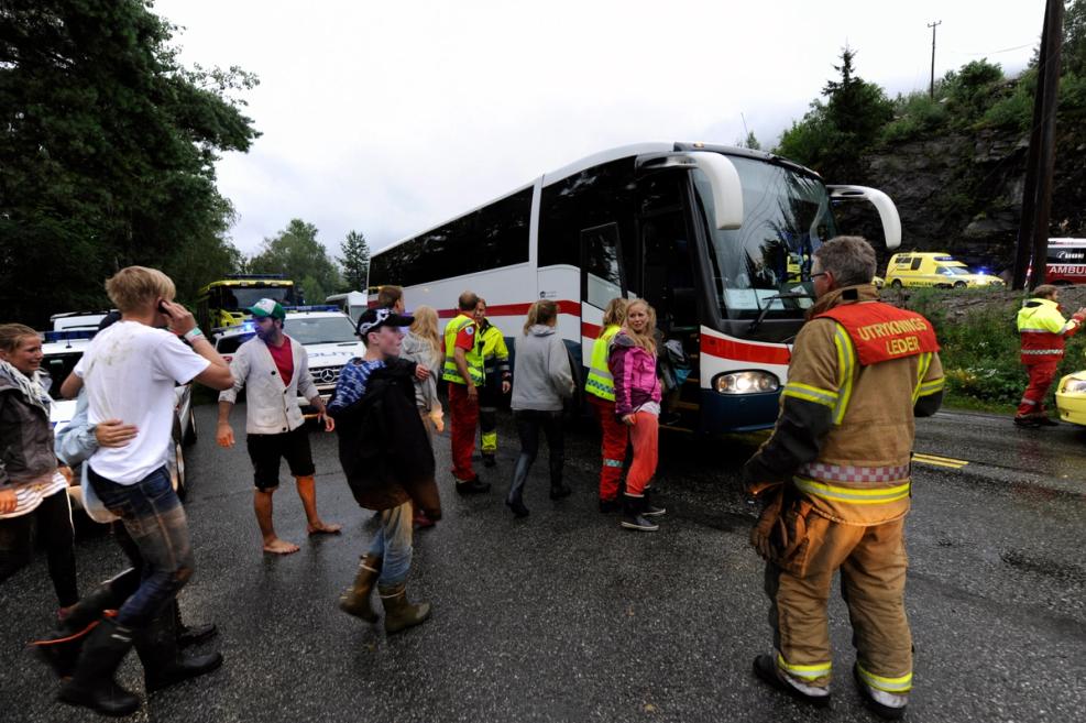 Foto. Unknown number of people in front of a bus. Wet asphalt. Trees and gray sky in the background.