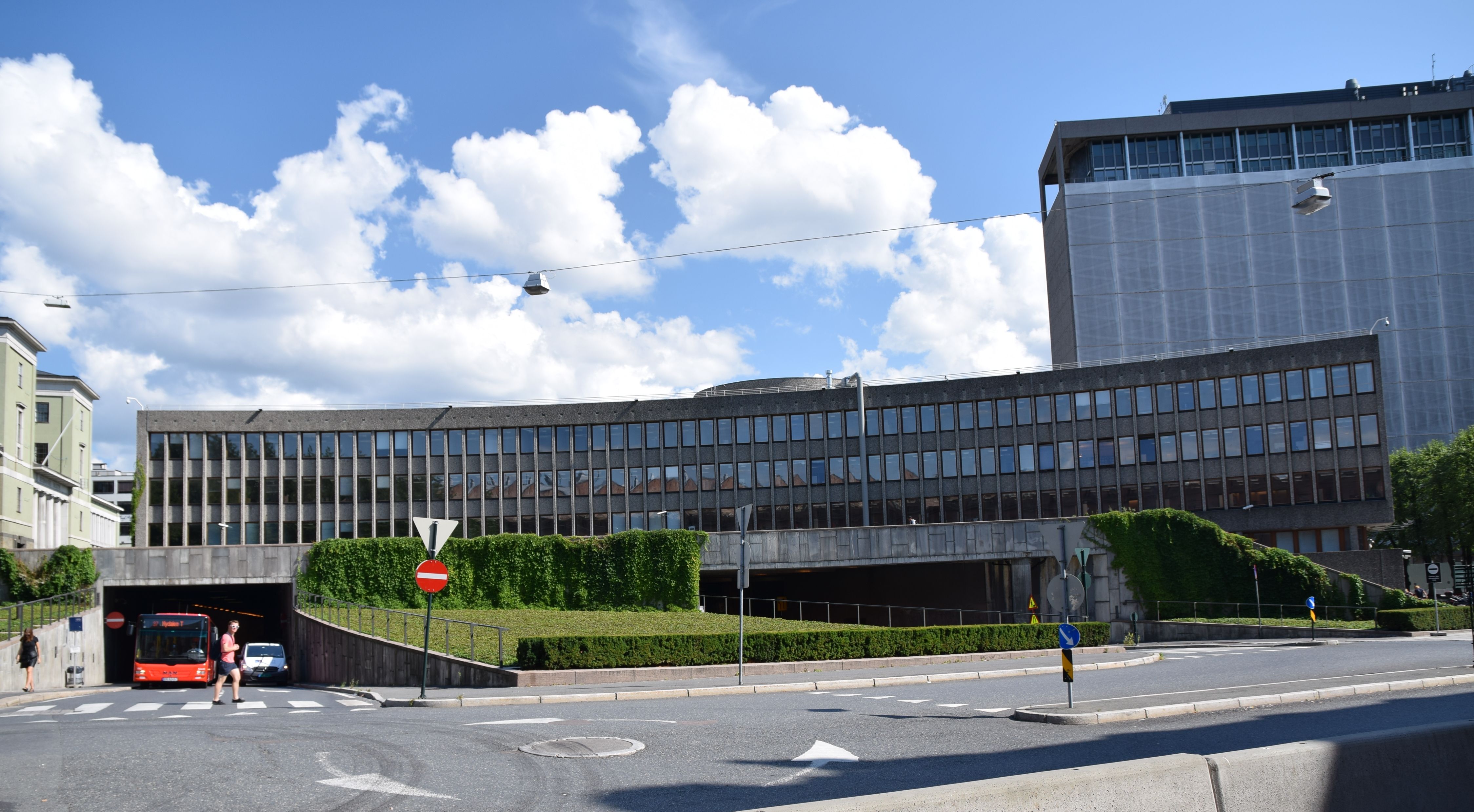 A low oblong office building. One person on the pavement, and one on the crosswalk. A red bus on its way out of the tunnel. the background a high-rise building.