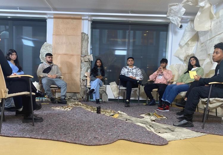 Photo. Nine people sitting in a circle on chairs. On the floor lies some sort of a grey carpet. On the wall to the right large sheets of paper are seemingly randomly placed. One of the persons are talking, the rest are observing that person.