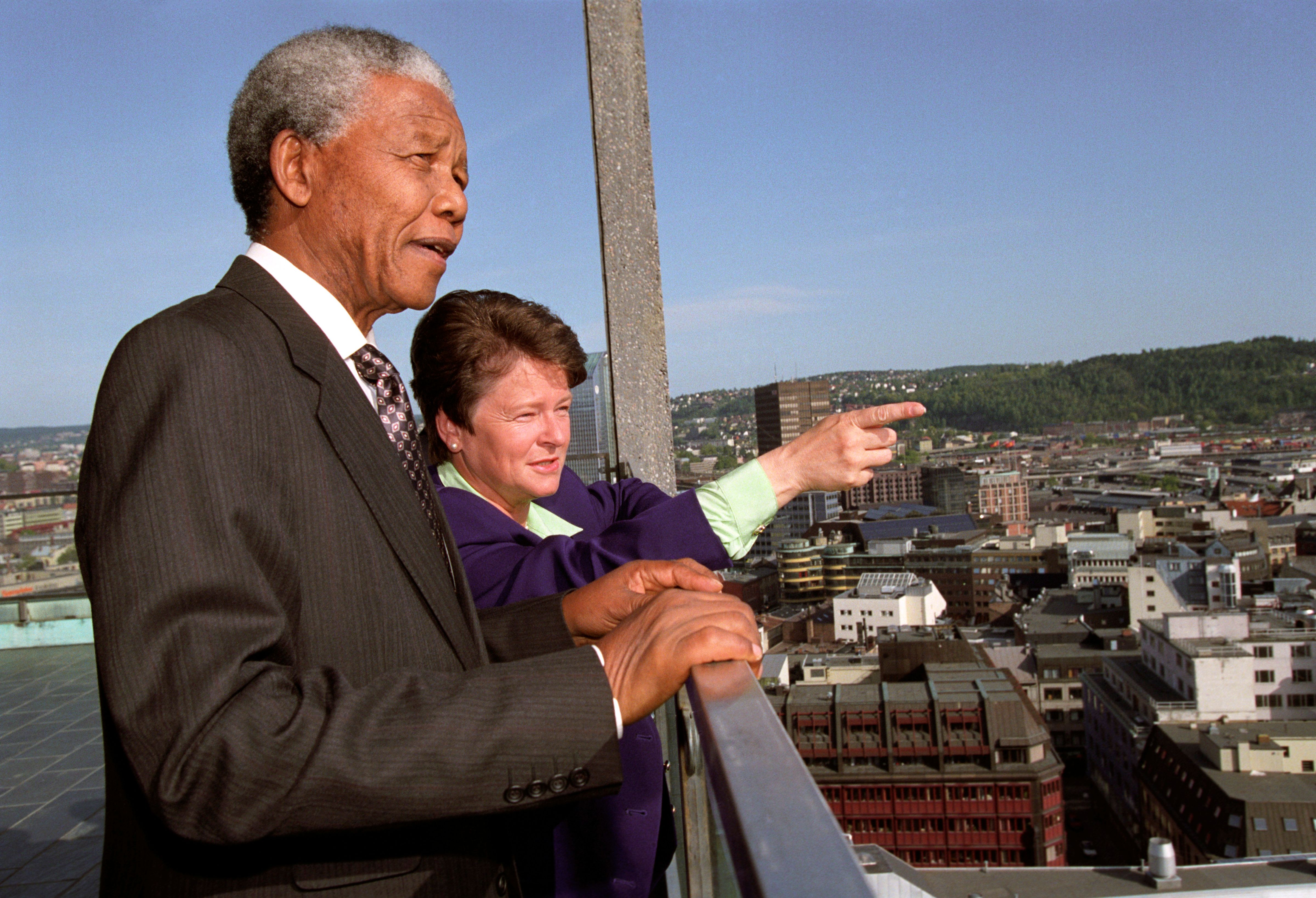 Two people in suits on a roof terrace. One points out towards the view. In the background cityscape.