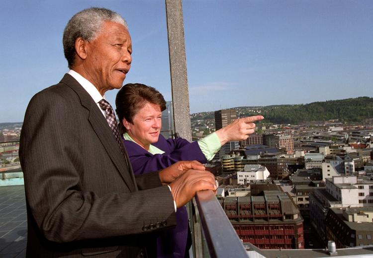 Two people in suits on a roof terrace. One points out towards the view. In the background cityscape.