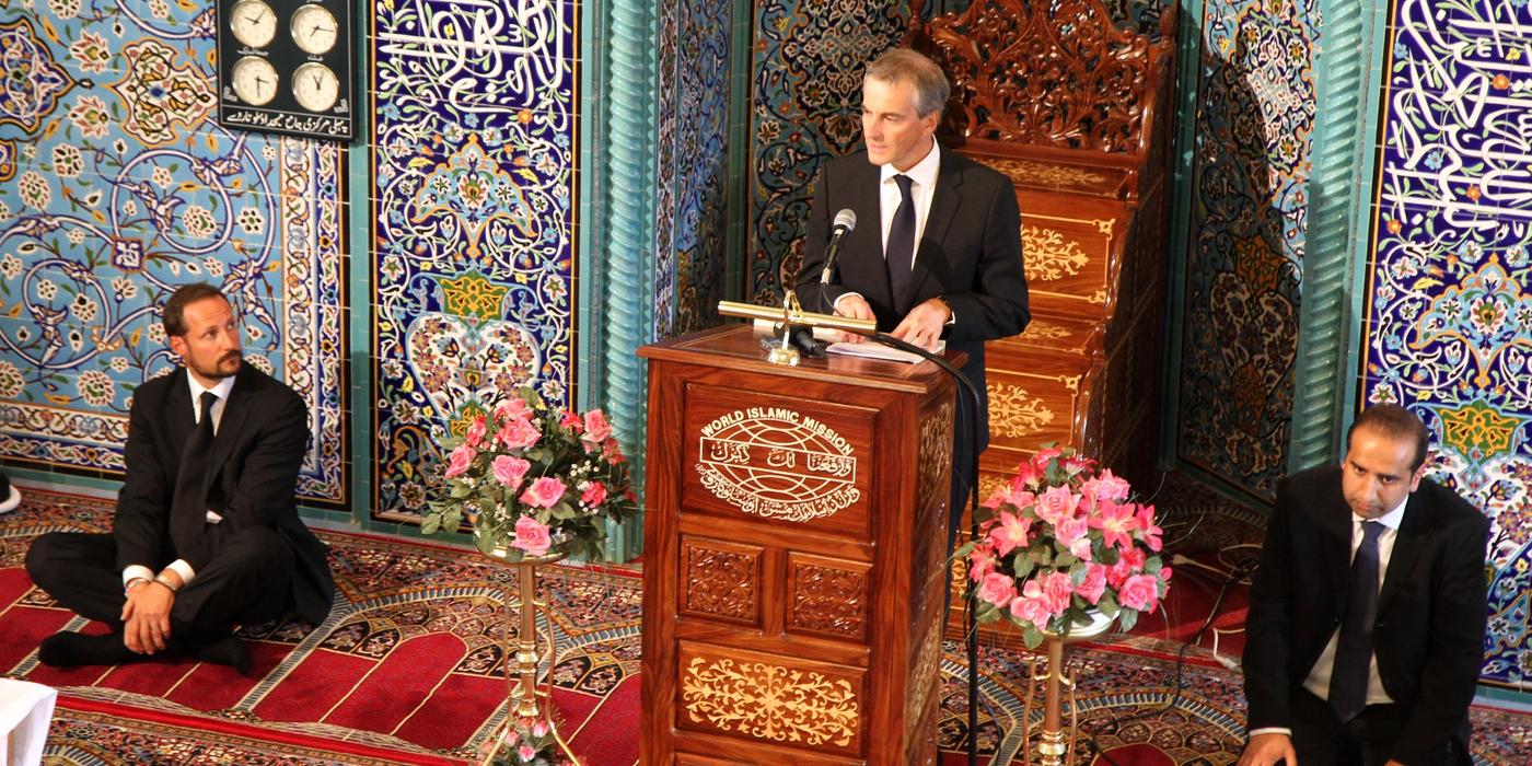 A person dressed in a suit on a lectern in front of a flower arrangement and a wall with art decoration. On either side of the lectern, a man in a suit sits on an ornate wall-to-wall carpet. On the wall hangs a picture frame with seven clocks showing different times.