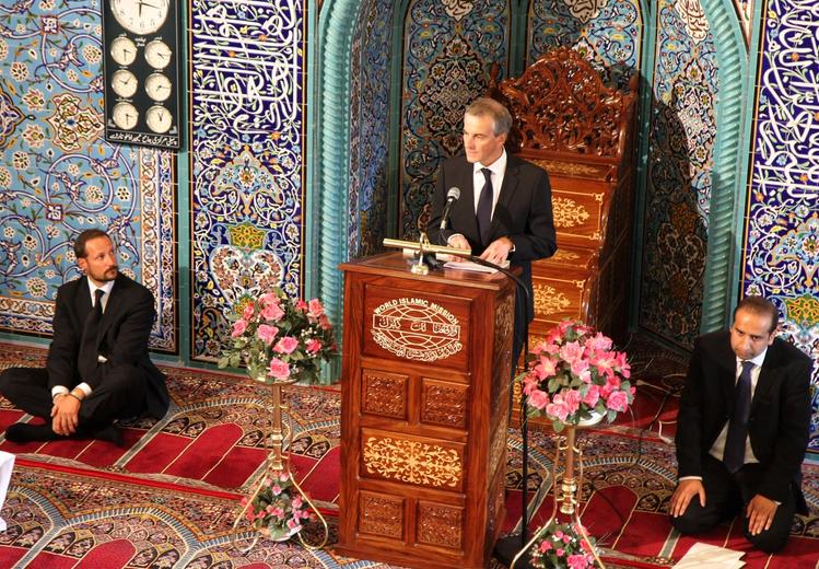 A person dressed in a suit on a lectern in front of a flower arrangement and a wall with art decoration. On either side of the lectern, a man in a suit sits on an ornate wall-to-wall carpet. On the wall hangs a picture frame with seven clocks showing different times.