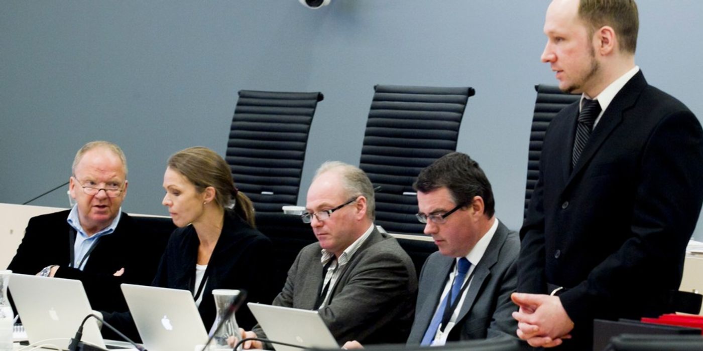 Fire mennesker sitter ned og ser frem for seg. Terroristen, Breivik, står oppreist til høyre i bildet. 
