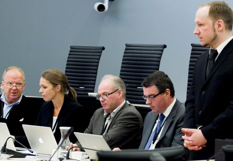 Fire mennesker sitter ned og ser frem for seg. Terroristen, Breivik, står oppreist til høyre i bildet. 