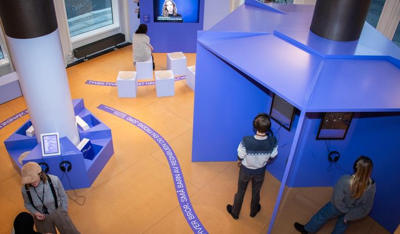 A photo taken from above. An exhibition with purple lines on the floor. People stand around and look at the exhibition. Various purple exhibition installations with screens display information.